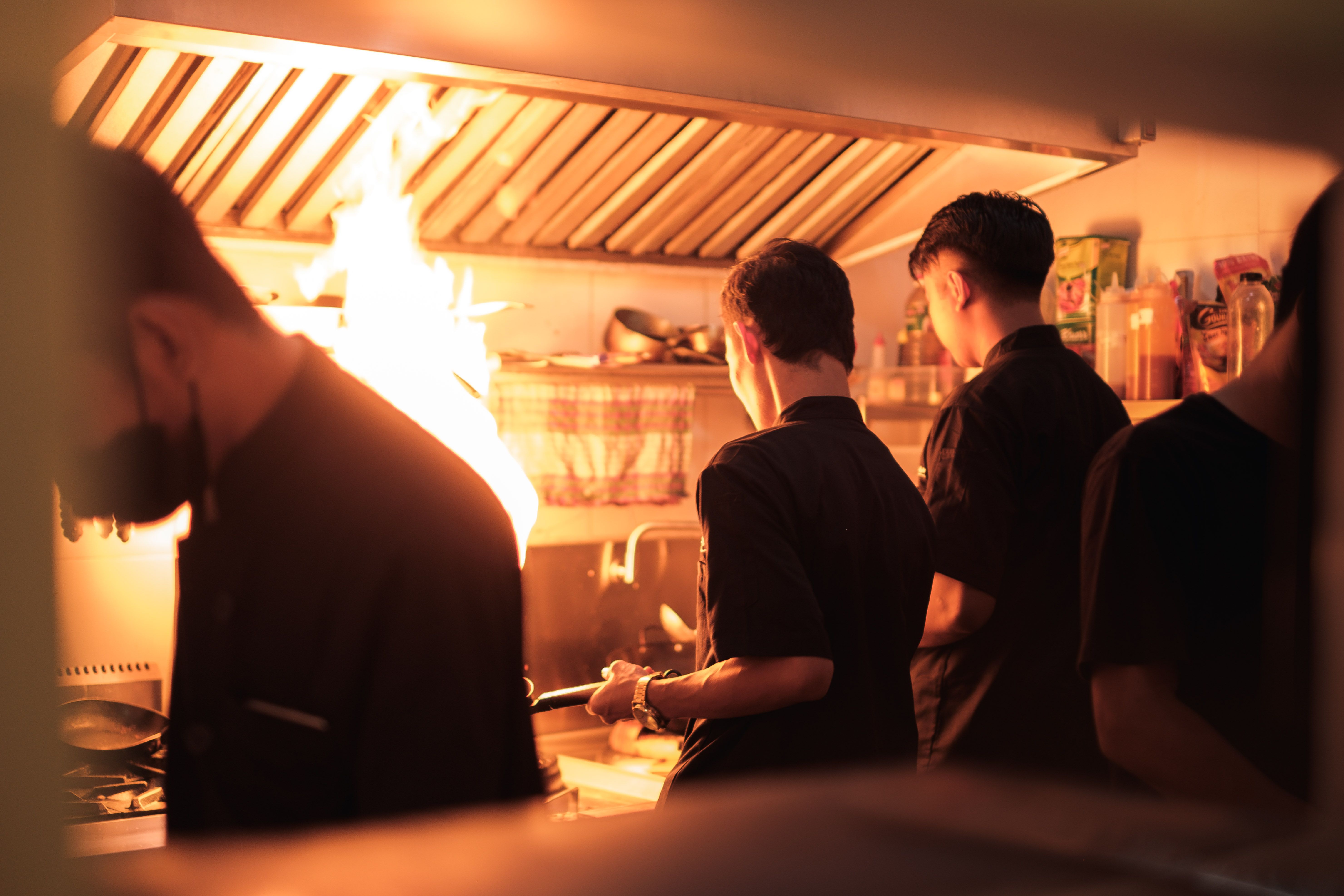 Brigade de cuisine au travail devant une flamme vive, sous une hotte d'extraction professionnelle.